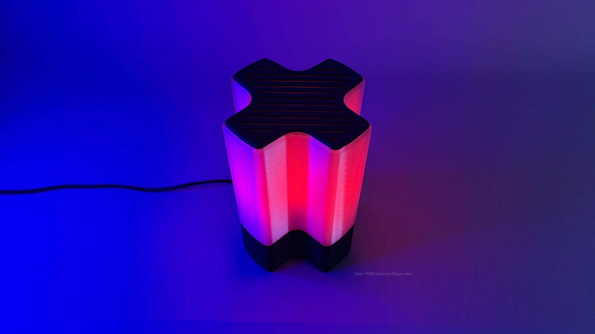 Front angled view of Nebu 3D printed modern tabletop lamp in clear shade, showing double-walled transparent design with lamp on in RGB mode